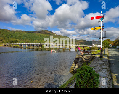 Adult male female relax on mawddach riverside, wooden white toll bridge and old railway signal in background, tree covered hills Stock Photo