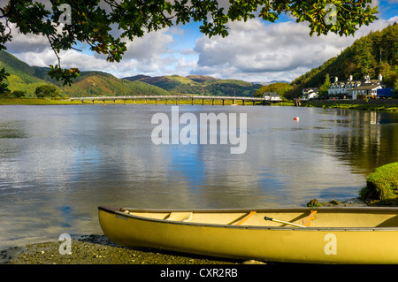 A yellow canoe in the foreground of the river mawddach with reflected blue sky, tree covered hills, white bridge in background