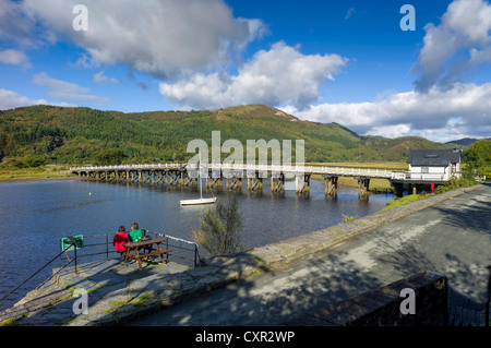 Adult male & female sitting at picnic table in foreground facing river mawddach with adjacent wooden toll bridge, forested hills Stock Photo