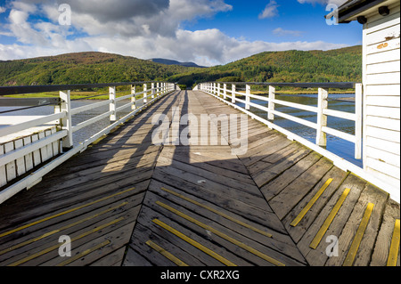 A view looking across an old wooden toll bridge, converging in the distance to a background of sunny tree covered hills Stock Photo