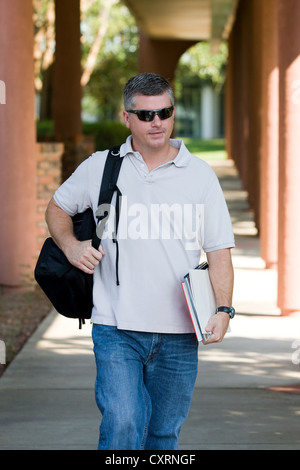 Mature adult student on campus walks to class carrying his books and backpack as he goes back to college for more education. Stock Photo