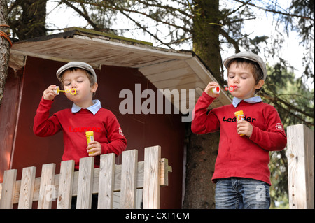 Twin boys, 4, wearing flat caps and blowing soap bubbles in a garden in front of a tree house Stock Photo