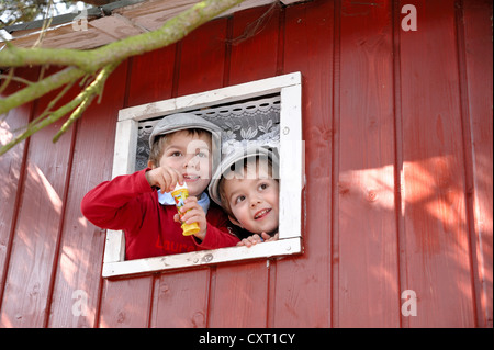 Twin boys, 4, wearing flat caps and blowing soap bubbles from the window of a tree house Stock Photo