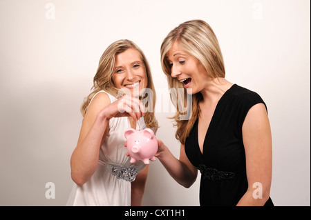 Twin sisters, one putting a 20 euro bill in the pink piggy bank of the other Stock Photo
