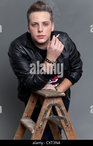 Young man in a leather jacket smoking a cigarette whilst resting on an old wooden ladder Stock Photo
