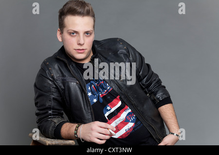 Young man smoking a cigarette wearing a leather jacket while leaning casually against an old wooden ladder Stock Photo