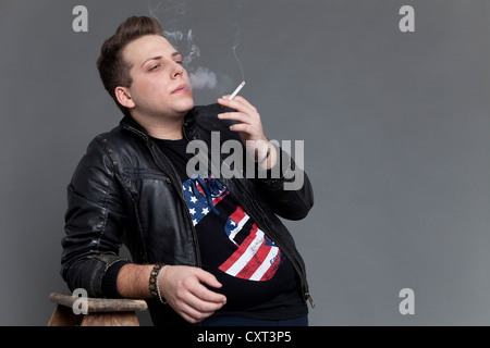 Young man smoking a cigarette wearing a leather jacket while leaning casually against an old wooden ladder Stock Photo