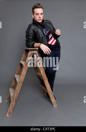 Young man with a cigarette wearing a leather jacket and jeans while leaning casually against an old wooden ladder Stock Photo