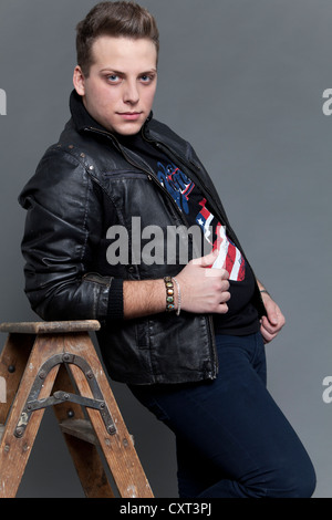 Young man wearing a leather jacket and jeans leaning casually against an old wooden ladder Stock Photo