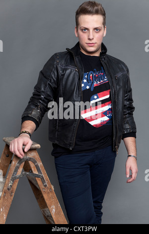 Young man wearing a leather jacket and jeans leaning against an old wooden ladder Stock Photo