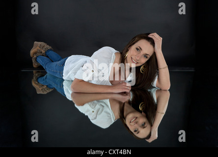 Young smiling woman wearing a white top and jeans, lying on a mirror, mirrored image Stock Photo