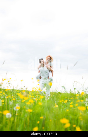 Young man carrying his girlfriend piggyback across a floral meadow Stock Photo