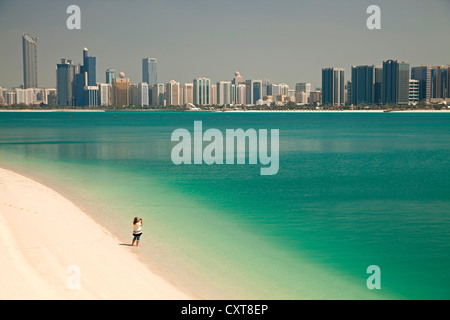 Individual tourist on the beach in front of the Heritage Village taking a photograph of the skyline of Abu Dhabi Stock Photo