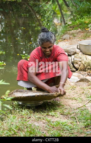 Vertical portrait of an Indian woman waterproofing a handmade grass basket with mud on the riverbank in Kerala.