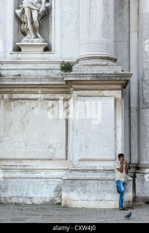 Young Asian man in jeans leaning against the Chiesa di San Stae in Venice, smoking a cigarette and speaking on his cellphone. Stock Photo