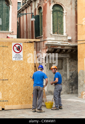 Two builders identically dressed in blue T-shrits, grey trousers and hard hats conferring about something on site in Venice Stock Photo