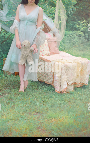 Cropped shot of a pale young woman wearing a fancy blue nightgown in the woods and holding a teddybear beside a child's bed