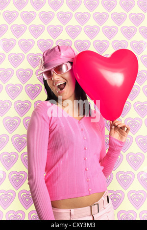 Young, happy woman wearing sunglasses and a pink cap, holding a heart-shaped balloon Stock Photo