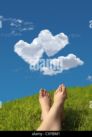 Woman lying on a meadow, detail of feet with a heart-shaped cloud formation in the sky Stock Photo