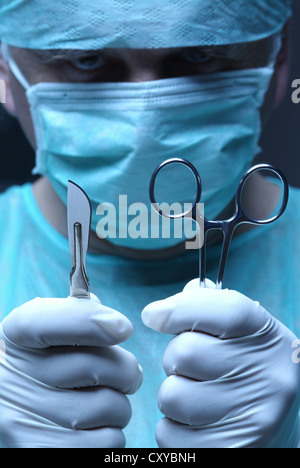 Surgeon wearing a face mask and holding a scalpel and a hemostatic clamp or arterial forceps in his hands during surgery Stock Photo