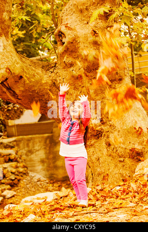 Picture of little cute girl play in autumn park, adorable sweet kid throwing up old dry leaves and laughing, nice small female Stock Photo