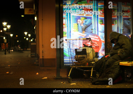 Man sleeping at a bus stop on a freezing cold winter's night in front of a poster of the Berlin City Mission calling for Stock Photo