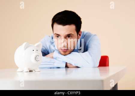 Young man sitting pensively in front of a piggy bank Stock Photo
