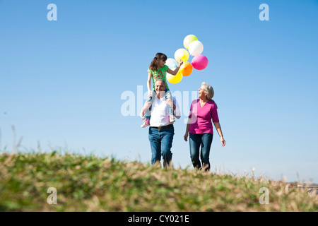 Girl holding many colourful balloons in her hand while sitting on her grandfather's shoulders, with her grandmother beside her Stock Photo
