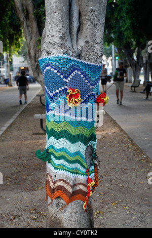 Colourful knitted and crocheted yarns wrap a trunk of a 'Yarn Bombed' tree which is a type of street art installation called Yarn bombing or yarnbombing in Rothschild street Tel Aviv Israel Stock Photo