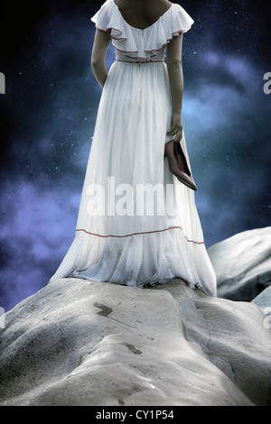 a woman in a white dress with polka dots is standing on rocks, holding her shoes in her hands Stock Photo