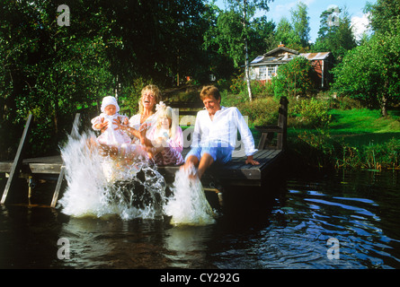 Family of four sitting barefoot on lake shore wooden dock kicking water into a sunny summer day in Sweden Stock Photo