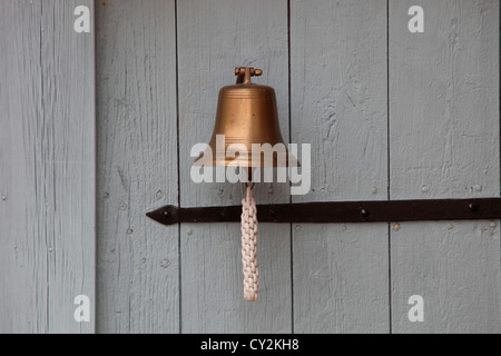 A small ship's bell with a knotted bell rope is hanging on a white wooden cottage door. Stock Photo