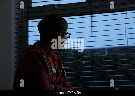 Young male wearing glasses, sitting in a dark room looking out of a window. Stock Photo
