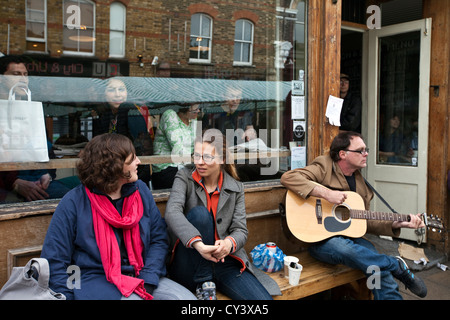 A man plays guitar outside a cafe in Broadway Market, London Fields, Hackney, London Stock Photo