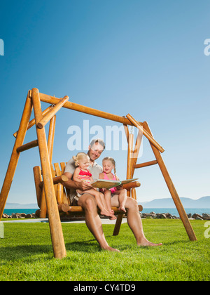 USA, Utah, Garden City, father with daughters (2-3, 4-5) on swing reading book Stock Photo