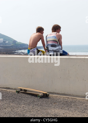 Young men with longboard skate board sit on wall overlooking beach. Westward Ho Devon. Stock Photo