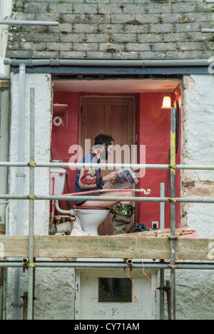 Builder taking a break whilst reading a newspaper on the toilet. Stock Photo