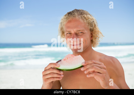 Smiling blonde man holding a piece of a watermelon while standing on the beach Stock Photo