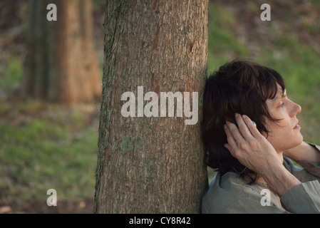 Young man leaning against tree trunk, listening to music with headphones, side view Stock Photo