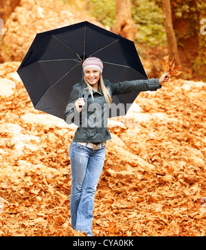 Photo of lovely woman walk under umbrella in rainy day in autumnal park, smiling girl hold in hand bouquet of orange dry leaves Stock Photo