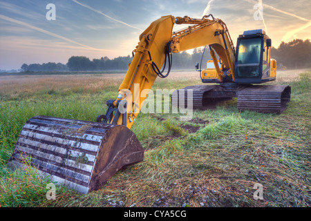 Yellow excavator in early morning light under a blue sky with condensation trails Stock Photo