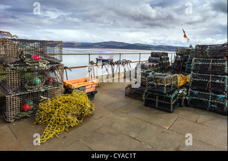 On the quayside is fishing gear comprised of crab and lobster pots, floats, ropes, nets , an inshore fishing boat lies at anchor Stock Photo