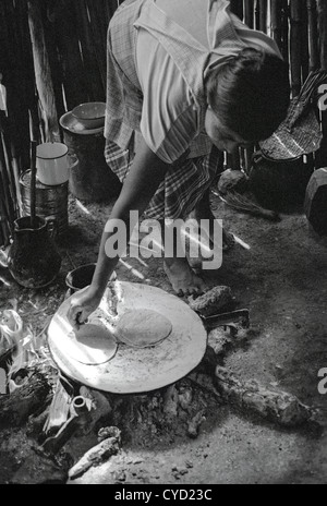 Black and white documentary photo of Kekchi woman cooking tortillas on a comal on an open fire in the floor of her house. Stock Photo