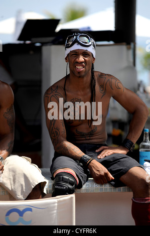 Young black guy with  upper body and arm tattoos sitting on bar wearing old motorcycle goggles and a knee pad Stock Photo
