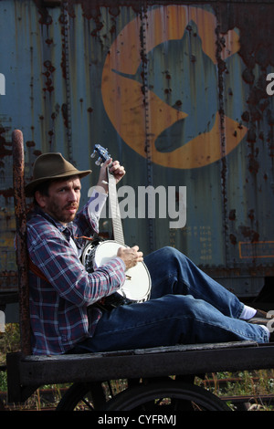 Male model as a hobo playing banjo while sitting on an old railroad baggage cart. Stock Photo