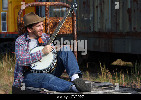 Male model as a hobo playing banjo while sitting on an old railroad baggage cart. Stock Photo