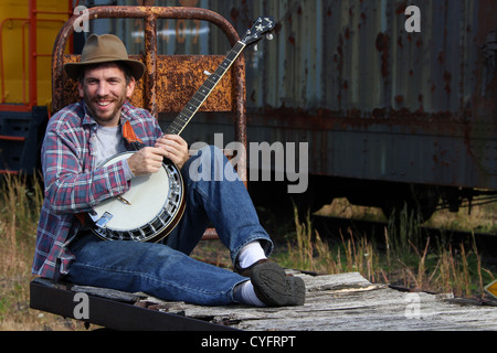 Male model as a hobo playing banjo while sitting on an old railroad baggage cart. Stock Photo