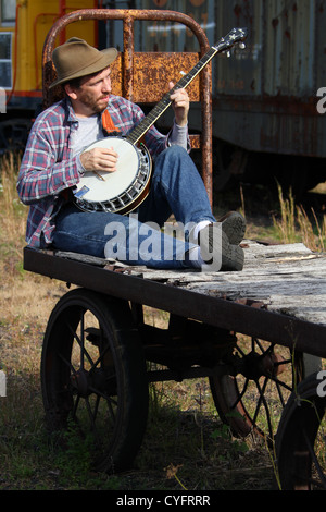 Male model as a hobo playing banjo while sitting on an old railroad baggage cart. Stock Photo