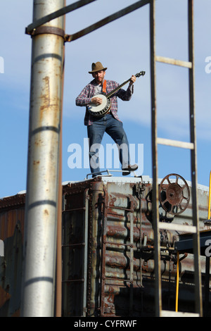 Male model as a hobo playing banjo while standing on a railroad box car. Stock Photo