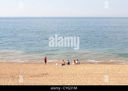 Group of young people sitting on UK beach with one holding a fishing rod Stock Photo